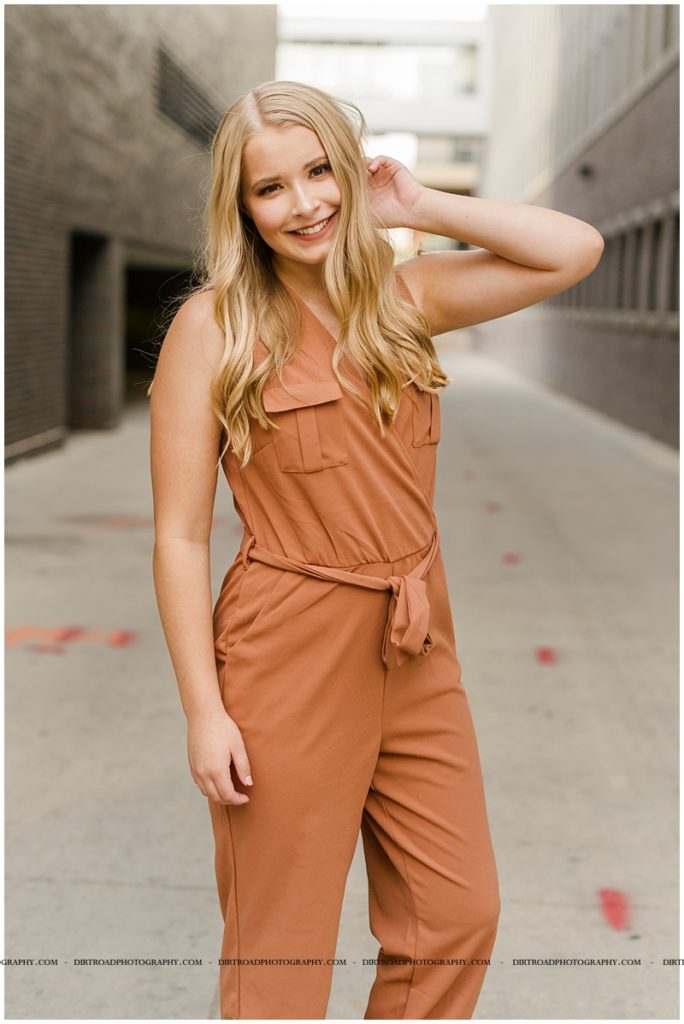 senior pictures of girl in downtown haymarket lincoln nebraska near the haymarket area. nebraska senior photographer. picture of girl wearing orange romper standing in alley surrounded by cement buildings with long blond curly hair.