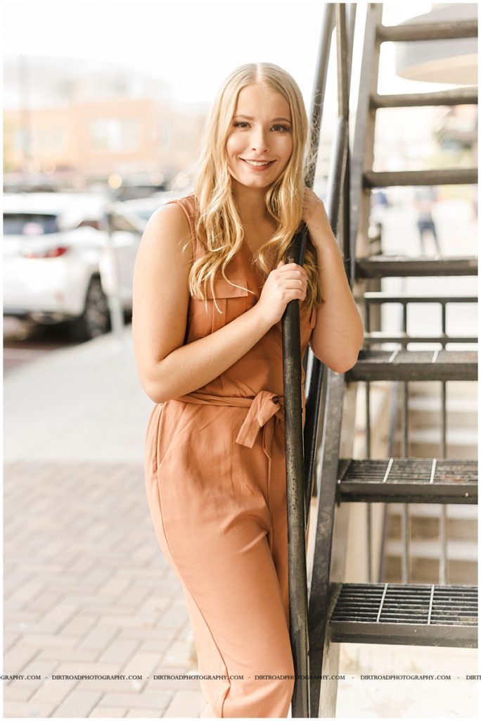 senior pictures of girl in downtown haymarket lincoln nebraska near the haymarket area. nebraska senior photographer. picture of girl wearing orange romper standing in alley surrounded by cement buildings with long blond curly hair.