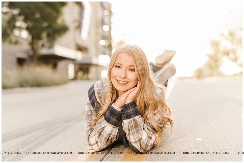 senior pictures of girl in downtown haymarket lincoln nebraska near the haymarket area. nebraska senior photographer. picture of girl wearing jeans with a plaid jacket standing in city street at sunset with long blond curly hair.