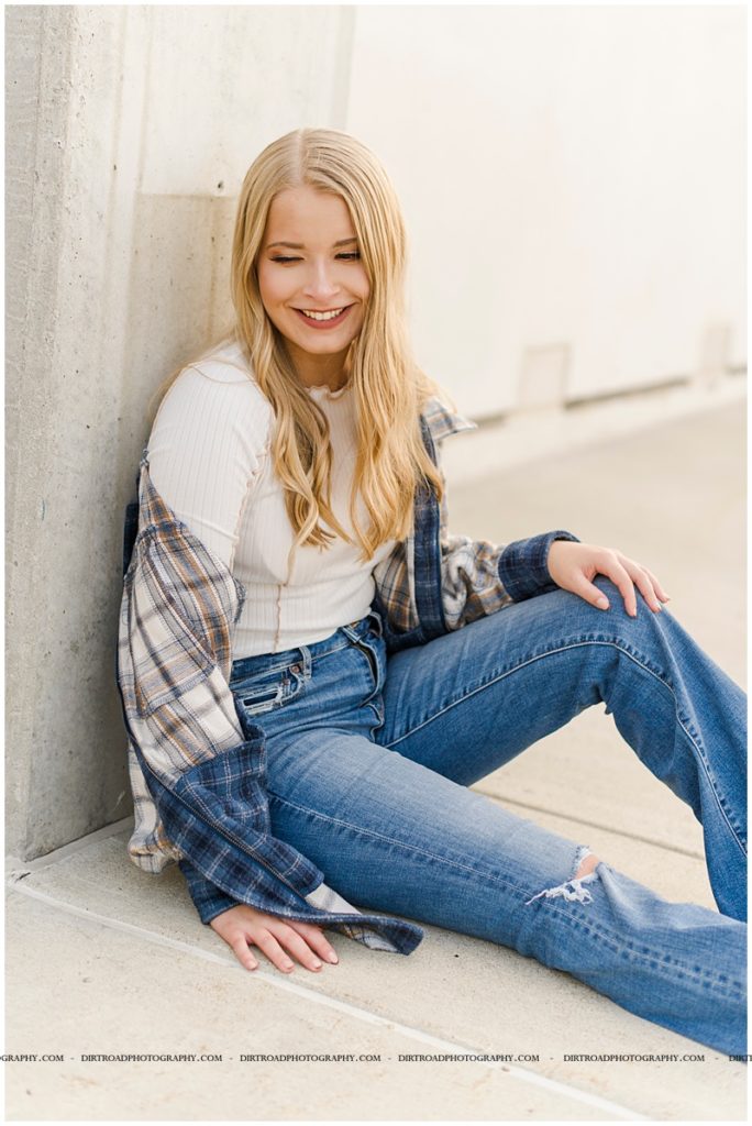 senior pictures of girl in downtown haymarket lincoln nebraska near the haymarket area. nebraska senior photographer. picture of girl wearing jeans with a plaid jacket standing in city street at sunset with long blond curly hair.