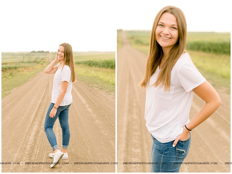 senior picture of girl wearing white tshirt with jeans and white tennis shoes with long brown hair. girl standing on dirt road at sunset with corn and big hill in the background. lincoln southeast high school. nebraska senior photographer located near lincoln, nebraska.