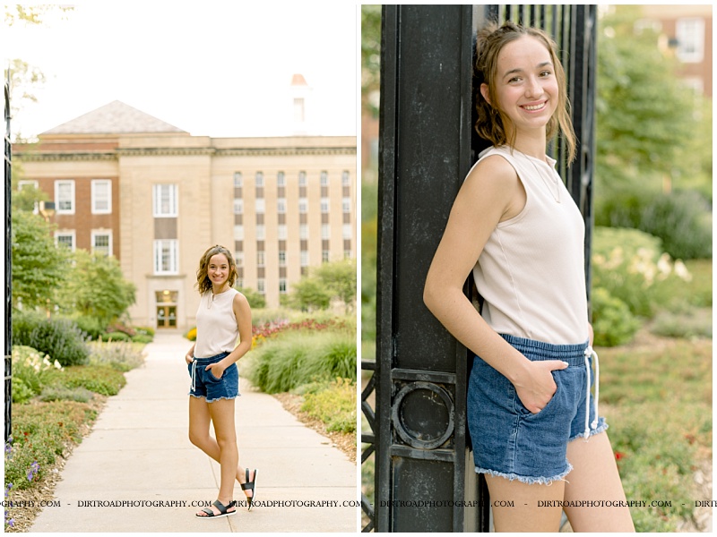 senior picture of girl standing on unl campus by big metal gate and with flowers in a walkway. senior girl is wearing jean shorts and tan tank top with hair in a ponytail. lincoln southwest high school. nebraska senior photographer located near lincoln, nebraska.