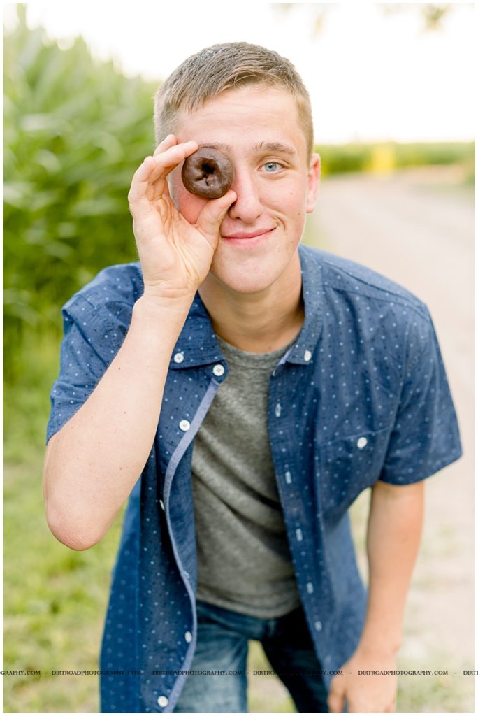 senior picture of boy with donuts. boy holding chocolate donut by his eye. milford nebraska high school. boy wearing blue button up shirt with jeans standing by road at sunset. nebraska senior photographer located near lincoln, nebraska.