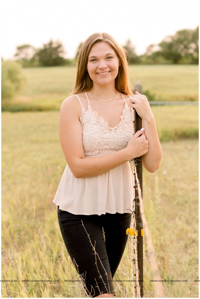 picture of senior girl taken at sunset. lincoln southeast high school. picture of girl with long brown hair wearing black distressed denim jeans wearing cream colored tank top with lace. standing next to fence post at sunset. nebraska senior photographer located near lincoln, nebraska.