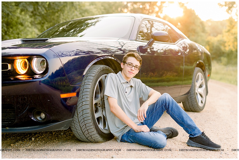 picture of senior boy sitting by dodge charger car. boy wearing button up gray shirt with jeans and black tennis shoes. picture of senior boy with blue car taken on gravel road at sunset. adams high school. nebraska senior photographer located near lincoln, nebraska.