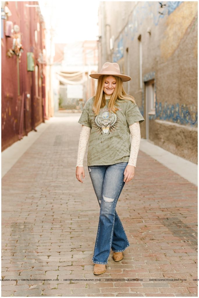 wilber-clatonia high school. senior picture of girl walking down brick alley wearing felt hat and flare jeans with graphic tee. boho senior picture style. nebraska senior photographer located near lincoln, nebraska.