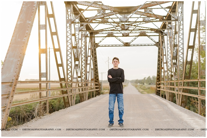 senior pictures of boy sitting standing on gravel road with cement bridge with metal overhang. senior boy wearing a black long sleeve shirt and jeans. senior pictures of ashland-greenwood high school senior. pictures taken by dirt road photography, lincoln nebraska best senior photographer.