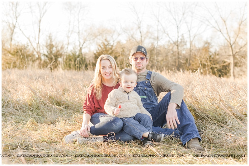 picture taken in fall with brown tall grass and pine trees. dad wearing denim overalls and boy wearing tan sweater with jeans. mom has long blond hair and is wearing red long sleeve shirt with jeans and brown boots. picture of parents holding boy at sunset. pictures taken by dirt road photography, lincoln nebraska best senior photographer.