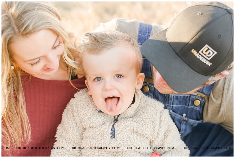 picture taken in fall with brown tall grass and pine trees. dad wearing denim overalls and boy wearing tan sweater with jeans. mom has long blond hair and is wearing red long sleeve shirt with jeans and brown boots. picture of parents holding boy at sunset. pictures taken by dirt road photography, lincoln nebraska best senior photographer.