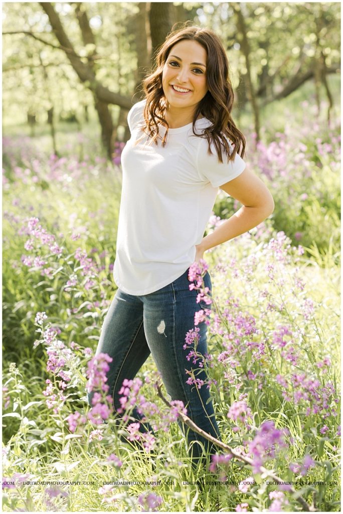picture of senior girl taken standing in tall purple flowers with trees near wilber, nebraska. senior picture of girl wearing white lace tank top with blue jeans with long blond hair standing in purple wildflowers. senior picture taken at sunset during the spring in nebraska. pictures taken by dirt road photography, lincoln nebraska best senior photographer.