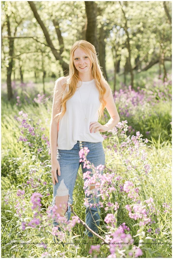 picture of senior girl taken standing in tall purple flowers with trees near wilber, nebraska. senior picture of girl wearing white lace tank top with blue jeans with long blond hair standing in purple wildflowers. senior picture taken at sunset during the spring in nebraska. pictures taken by dirt road photography, lincoln nebraska best senior photographer.
