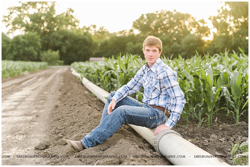 Luke sitting on a gravity flow irrigation pipe row with summer corn growing behind him at sunset for senior pictures in Nebraska.