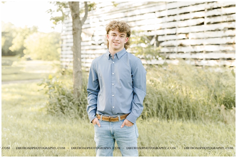 senior pictures of boy standing near grain bins at sunset wearing a dark red long sleeve shirt and jeans with cowboy boots. senior pictures of elkhorn south high school senior. pictures taken by dirt road photography, omaha nebraska best senior photographer.