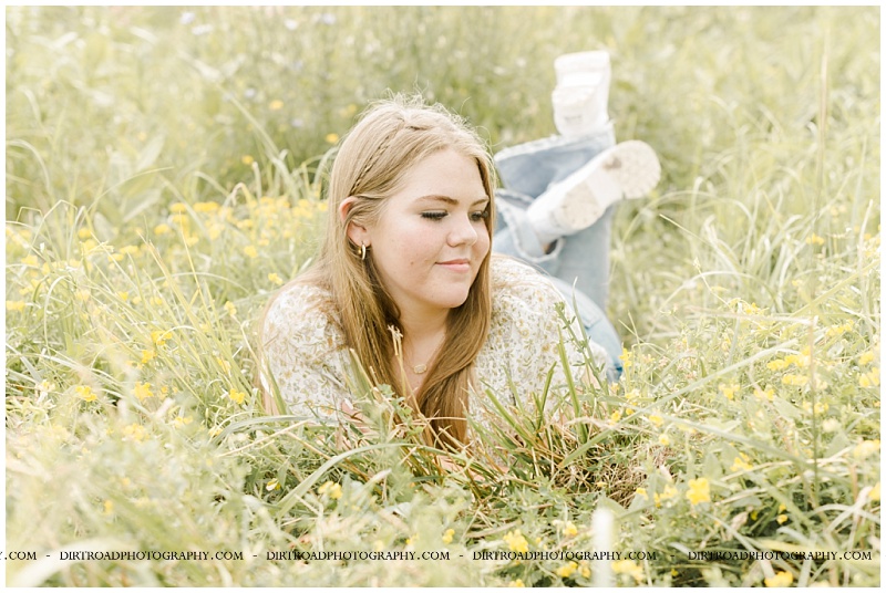 senior pictures of bella taken standing and sitting in nature with pretty wild flowers, tall grass and trees at sunset. senior girl is wearing american eagle denim flare jeans and floral tank top with long blond hair in two mini braids. Bella took senior pictures in the late summer in southeast nebraska. pictures taken by dirt road photography, hastings nebraska best senior photographer.