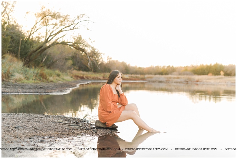 Papillion LaVista South high school senior pictures of girl in orange flowy dress sitting on rock of a pond in the fall at sunset. senior girl pictures with water. nebraska senior photographer located near lincoln, nebraska.